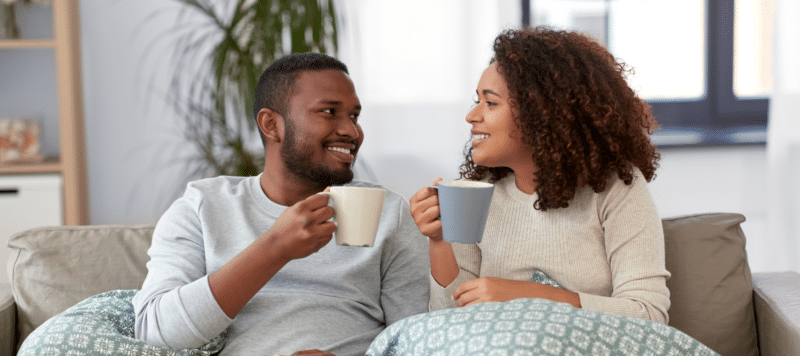 young couple sitting on a couch smiling at each other while drinking coffee